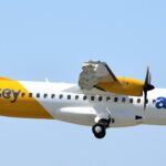 An Aurigny airlines aircraft is pictured in flight against pale blue sky. The aircraft is pictured from the side and appears to be at a slight angle descending, with landing gear out and the nose positioned lower right and the tail positioned upper left. The aircraft is mostly white with a deep yellow backend, tail, and engine. Dark blue branding can be seen along the side of the fuselage.
