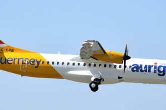 An Aurigny airlines aircraft is pictured in flight against pale blue sky. The aircraft is pictured from the side and appears to be at a slight angle descending, with landing gear out and the nose positioned lower right and the tail positioned upper left. The aircraft is mostly white with a deep yellow backend, tail, and engine. Dark blue branding can be seen along the side of the fuselage.