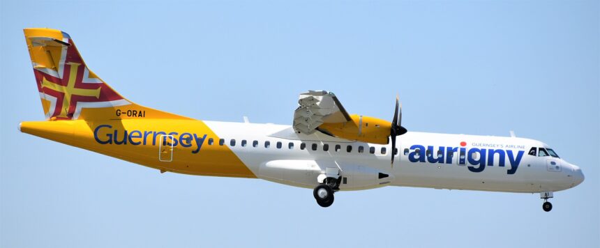 An Aurigny airlines aircraft is pictured in flight against pale blue sky. The aircraft is pictured from the side and appears to be at a slight angle descending, with landing gear out and the nose positioned lower right and the tail positioned upper left. The aircraft is mostly white with a deep yellow backend, tail, and engine. Dark blue branding can be seen along the side of the fuselage.