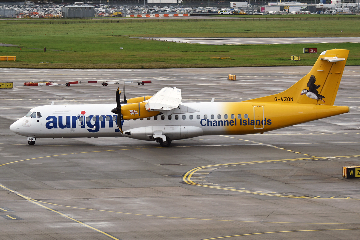 An Aurigny airlines aircraft is pictured on a taxiway or a runway at an airport. The aircraft is centred in the image, pictured from the side with the nose facing left and tail facing right. It is mostly white with deep yolk-yellow engines and back end, with an image of a bird on the tail. Navy blue branding can be seen along the side of the fuselage. in the foreground and midground the tarmac of the taxiway/runway can be seen, and the in the background a carpark can be seen just beyond a large patch of green grass.