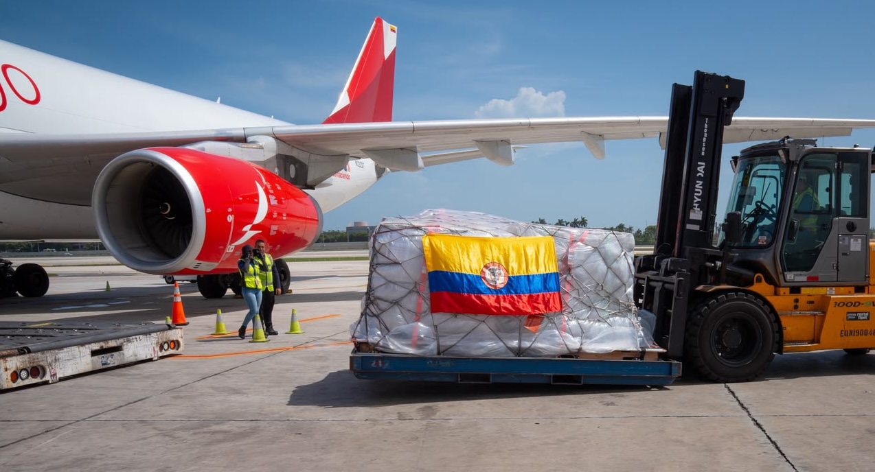 A yellow forklift on a tarmac positions a large, cargo-netted pallet wrapped in plastic next to an Avianca aircraft. A Colombian flag is draped across the front of the cargo pallet, while ground crew workers in high-visibility vests stand nearby under a clear sky.