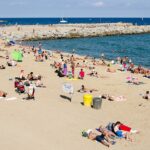 Image shows Barcelona beach on a sunny day with tourists on the sand and in the sea