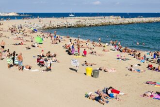 Image shows Barcelona beach on a sunny day with tourists on the sand and in the sea