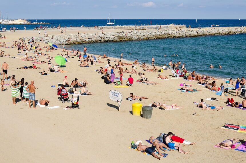 Image shows Barcelona beach on a sunny day with tourists on the sand and in the sea