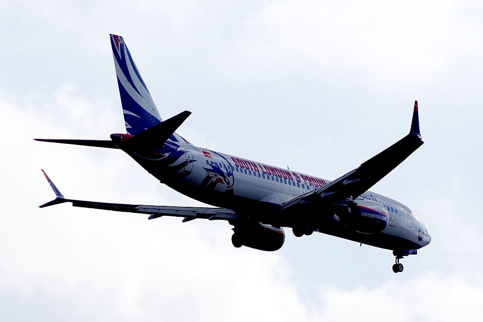 Image depicting a sunexpress.com Boeing 737 MAX landing against a cloudy grey sky. The aircraft is decorated with red, white and blue and an eagle is depicted on the side.