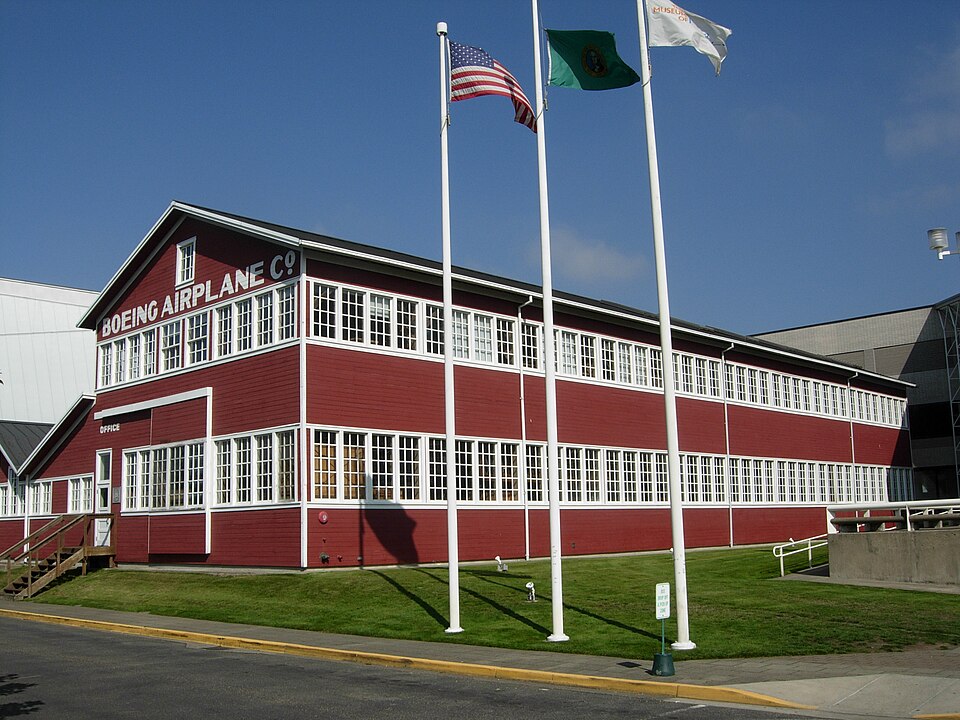 Picture depicting the Boeing Red Barn, Boeing's original manufacturing plant, which is now located at the Museum of Flight in Tukwila/Seattle, Washington. Three flags, including the U.S. flag, are flying out front of the barn, against a clear blue sky.