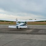 A small white Cessna 172 aircraft is pictured from behind at Strausberg airfield. The aircraft is white with blue strips along the main body. The foreground of the image is grey tarmac, the background is cloudy grey sky.
