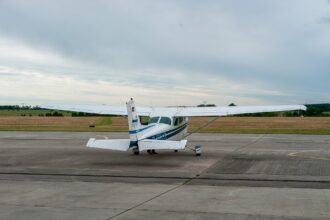 A small white Cessna 172 aircraft is pictured from behind at Strausberg airfield. The aircraft is white with blue strips along the main body. The foreground of the image is grey tarmac, the background is cloudy grey sky.