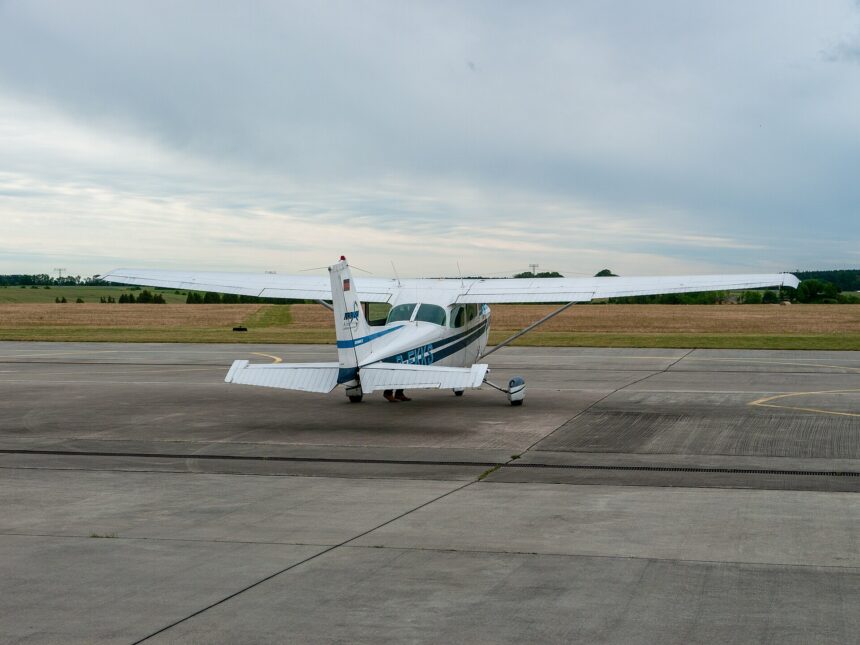 A small white Cessna 172 aircraft is pictured from behind at Strausberg airfield. The aircraft is white with blue strips along the main body. The foreground of the image is grey tarmac, the background is cloudy grey sky.