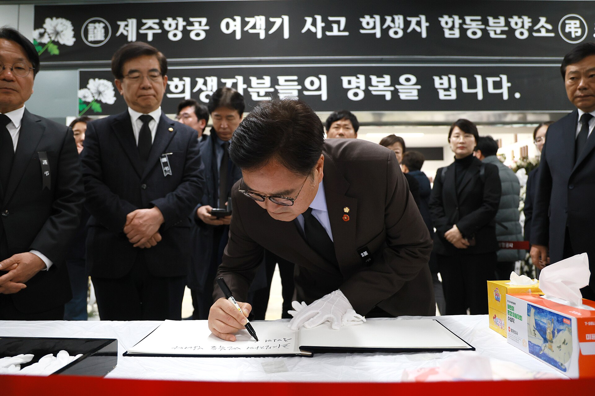 Chairman Woo Won-sik in Muan where Jeju Air Flight 2216 crashed on 2 January 2025. A group of well dressed East Asian people in suits gather around the suited chairman, who is bent over a table writing something down. In the background, black banners with Korean writing in white can be seen. 