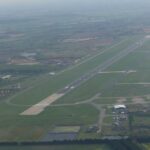 An aerial photograph of Doncaster Sheffield Airport. The runway can be seen extending from the centre, surrounded by grass. Airport buildings can be seen a short way away from the runway, and residential areas in the distance.