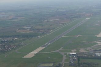 An aerial photograph of Doncaster Sheffield Airport. The runway can be seen extending from the centre, surrounded by grass. Airport buildings can be seen a short way away from the runway, and residential areas in the distance.