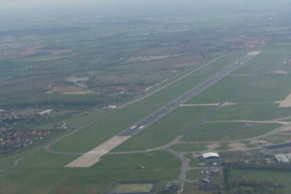 An aerial photograph of Doncaster Sheffield Airport. The runway can be seen extending from the centre, surrounded by grass. Airport buildings can be seen a short way away from the runway, and residential areas in the distance.