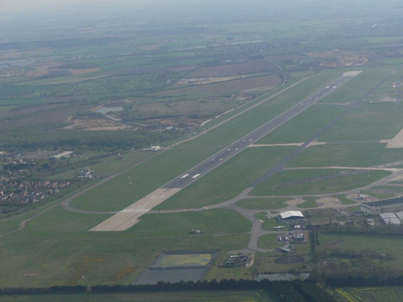 An aerial photograph of Doncaster Sheffield Airport. The runway can be seen extending from the centre, surrounded by grass. Airport buildings can be seen a short way away from the runway, and residential areas in the distance.