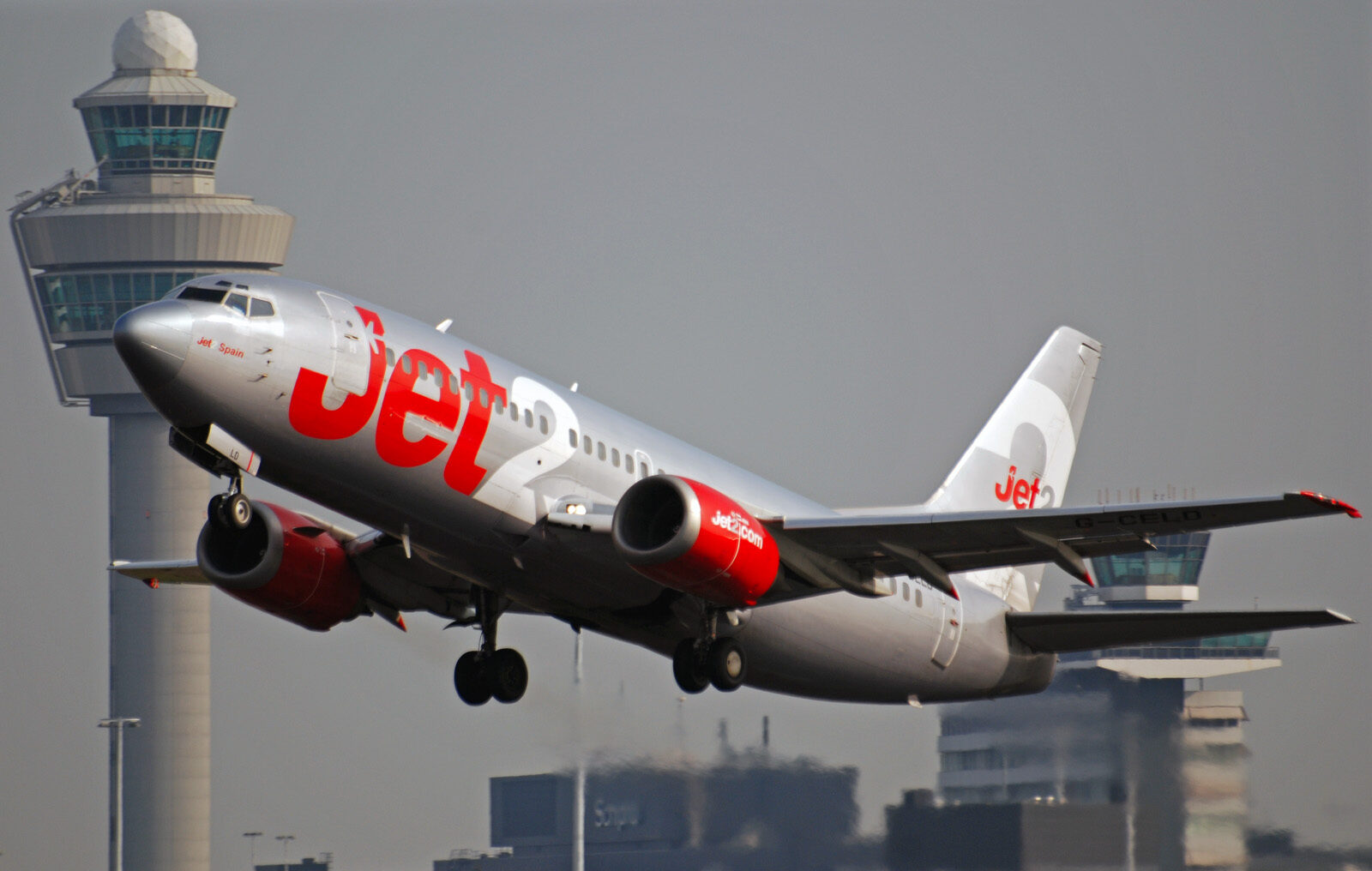 A Jet2 plane is taking off from an airport. The aircraft is grey with red and white 'Jet2' branding along the side of the fuselage and on the tail. The plane is pictured from the front at an upwards angle, and is positioned with the nose to the upper left and the tail to the lower right. In the background, various airport buildings can be seen, including a control tower. The sky in the background is grey.