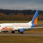 A Jet2 aircraft is pictured on a runway or taxiway at an airport. The aircraft is pictured from the front at an angle, with the nose to the centre right and the tail to the centre left. The plane is white, with a light blue tail with an orange and yellow sun logo on it, and 'Jet2holidays' branding along the lower front of the aircraft. In the foreground, tarmac separated by grass verges can be seen. In the middle distance, there is a row of trees and shrubbery, before a strip of grey blue sky in the background.