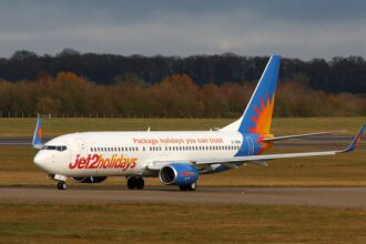 A Jet2 aircraft is pictured on a runway or taxiway at an airport. The aircraft is pictured from the front at an angle, with the nose to the centre right and the tail to the centre left. The plane is white, with a light blue tail with an orange and yellow sun logo on it, and 'Jet2holidays' branding along the lower front of the aircraft. In the foreground, tarmac separated by grass verges can be seen. In the middle distance, there is a row of trees and shrubbery, before a strip of grey blue sky in the background.