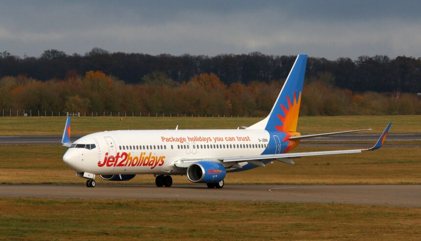 A Jet2 aircraft is pictured on a runway or taxiway at an airport. The aircraft is pictured from the front at an angle, with the nose to the centre right and the tail to the centre left. The plane is white, with a light blue tail with an orange and yellow sun logo on it, and 'Jet2holidays' branding along the lower front of the aircraft. In the foreground, tarmac separated by grass verges can be seen. In the middle distance, there is a row of trees and shrubbery, before a strip of grey blue sky in the background.