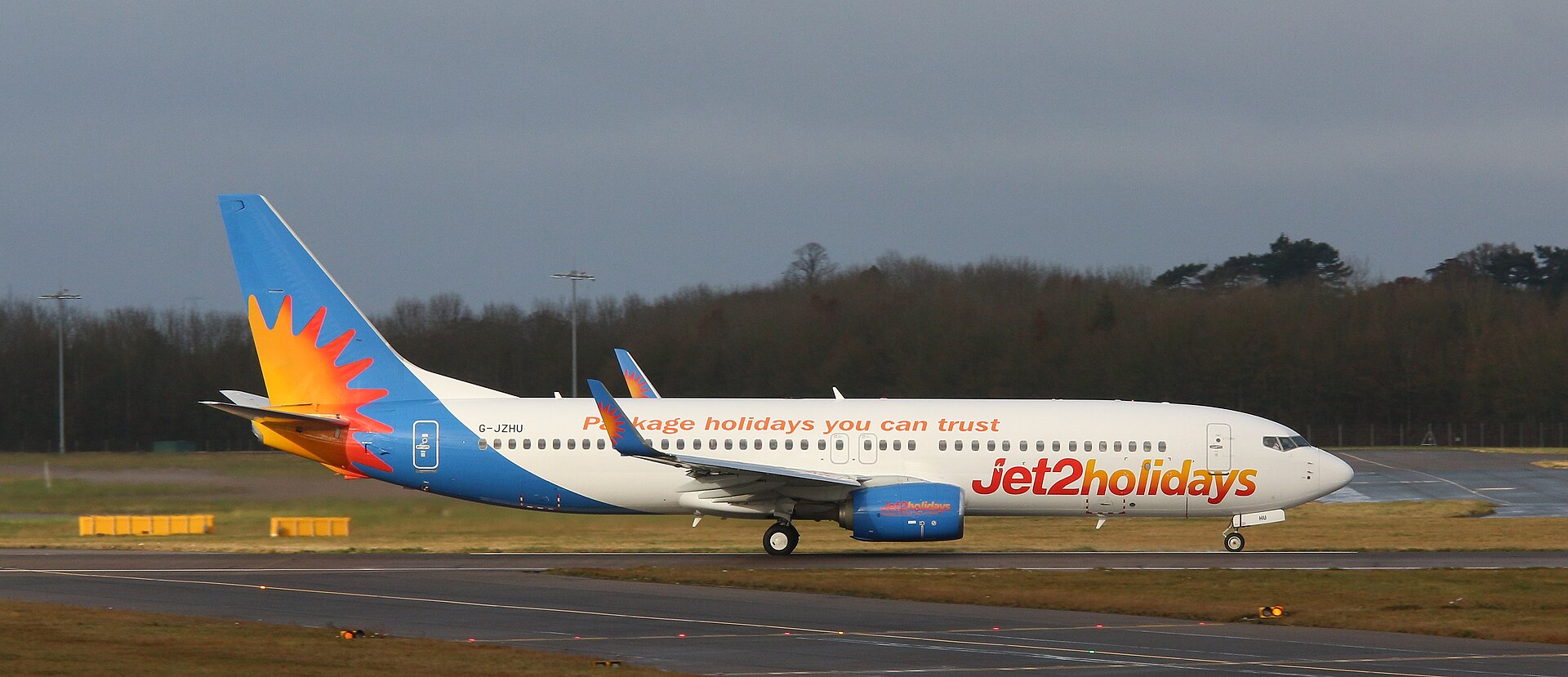 A Jet2 aircraft is pictured on a runway or taxiway at an airport. The aircraft, which is pictured from the side, is white, with a light blue tail with an orange and yellow sun logo on the left, and 'Jet2holidays' branding along the lower front of the aircraft, which is to the right. In the foreground, tarmac separated by grass verges can be seen. In the middle distance, there is a row of trees and shrubbery, before a strip of grey blue sky in the background.