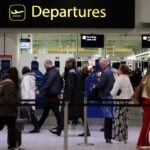 Passengers queue to enter security below a yellow Departures sign at Gatwick Airport.