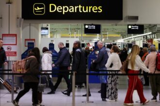 Passengers queue to enter security below a yellow Departures sign at Gatwick Airport.