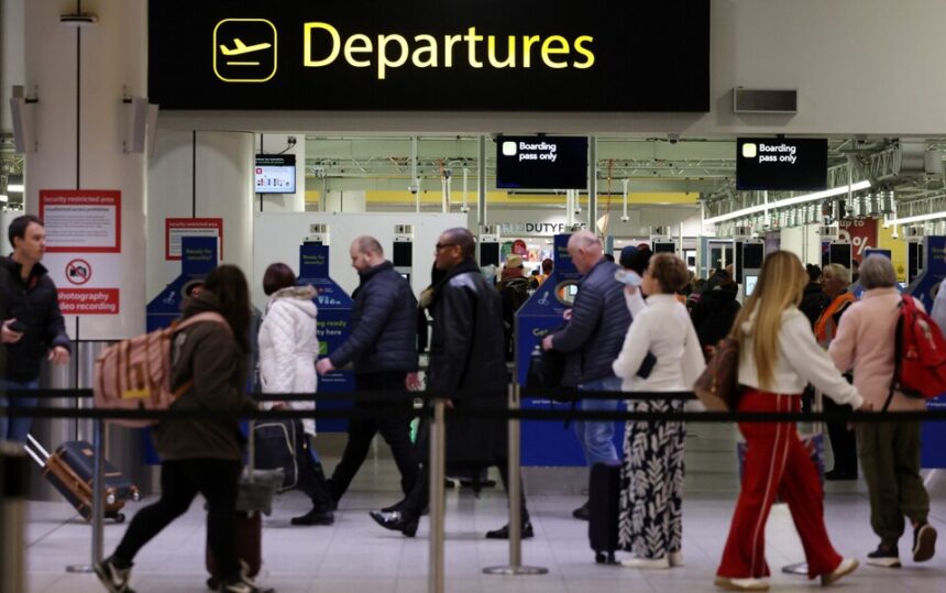 Passengers queue to enter security below a yellow Departures sign at Gatwick Airport.