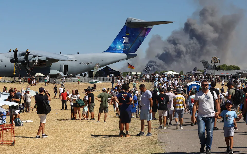 Spectators at the Athens Flying Week airshow are pictured walking away from the event, with smoke coming from the crash site filling the area behind them.