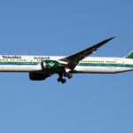 A Saudia Airlines plane is pictured in flight. The aircraft is mostly white with green branding on the tail and engine. The name of the airline is written across the front end fuselage in the same green, and a green stripe along the centre of the fuselage can be seen across the whole aircraft as well. The nose of the aircraft faces left and the tail faces the right. The background is a bright blue cloudless sky.