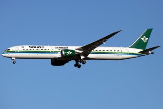 A Saudia Airlines plane is pictured in flight. The aircraft is mostly white with green branding on the tail and engine. The name of the airline is written across the front end fuselage in the same green, and a green stripe along the centre of the fuselage can be seen across the whole aircraft as well. The nose of the aircraft faces left and the tail faces the right. The background is a bright blue cloudless sky.
