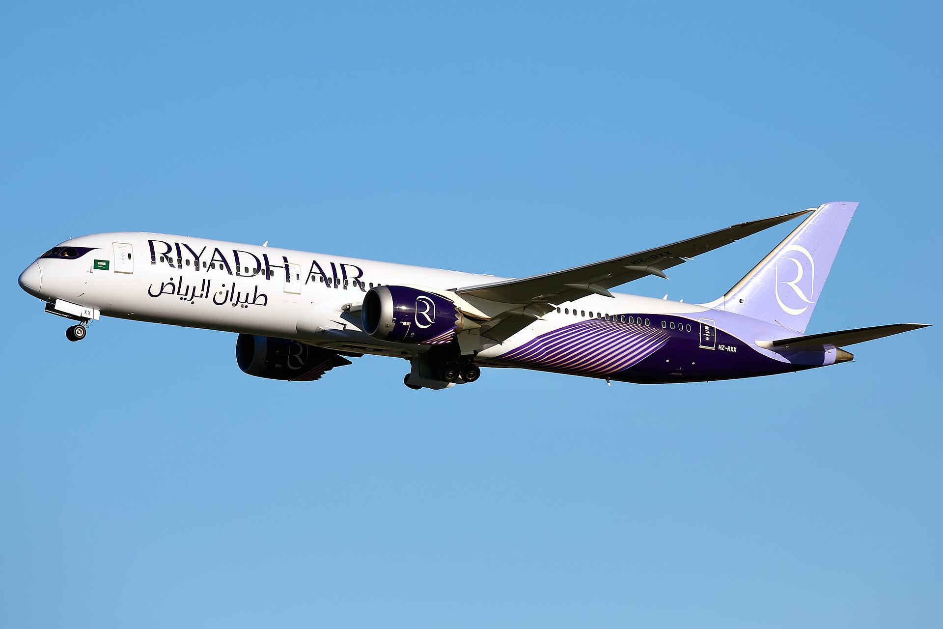 A Riyadh Air plane in flight. The aircraft is mostly white with purple branding on the tail, backend, and engine. The name of the airline is written across the front end fuselage in purple as well. The nose of the aircraft faces left and the tail faces the right. The aircraft is pictured at a small angle, making the nose marginally higher than the tail. The background is a bright blue cloudless sky. 