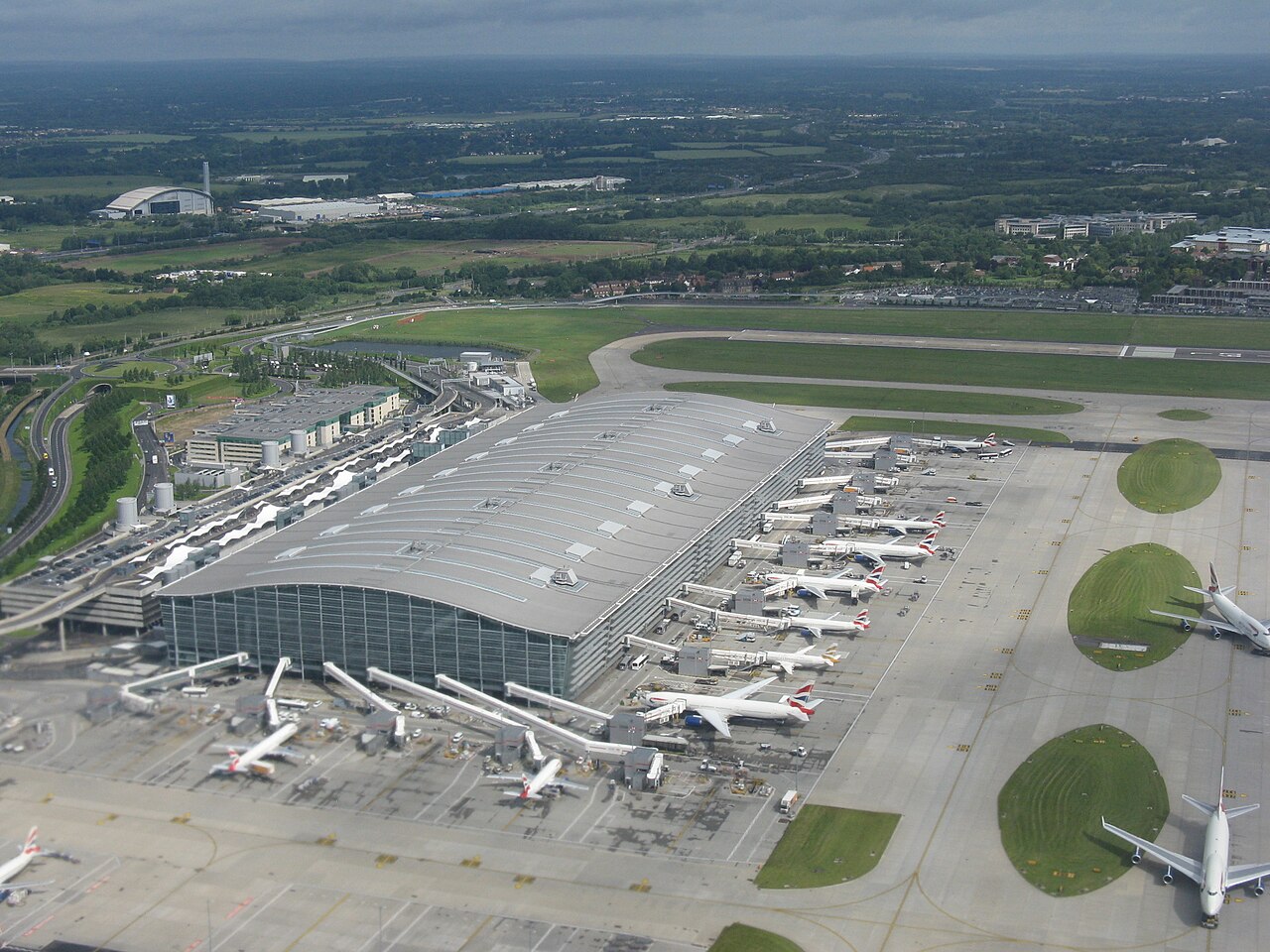 An aerial view of Heathrow Airport's Terminal 5 with planes stationed outside of its big glass exterior. The green grass of the runway is visible, with other small buildings scattered around.