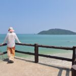 Image shows a person standing at a balcony overlooking a beach in Thailand on a clear day, with mountains in the background