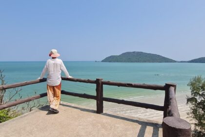 Image shows a person standing at a balcony overlooking a beach in Thailand on a clear day, with mountains in the background