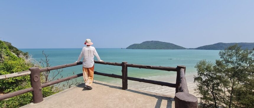 Image shows a person standing at a balcony overlooking a beach in Thailand on a clear day, with mountains in the background