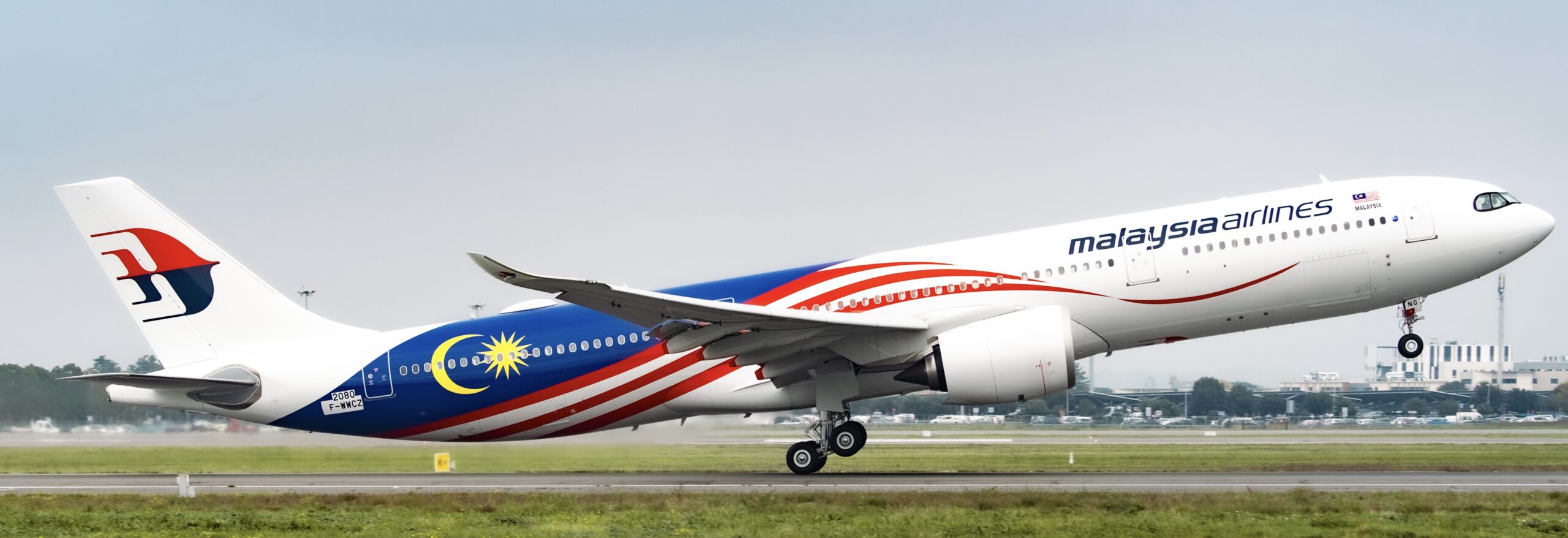 A red, white, and blue Malaysia Airlines jet taking off from a runway. The runway is between two grassy verges.