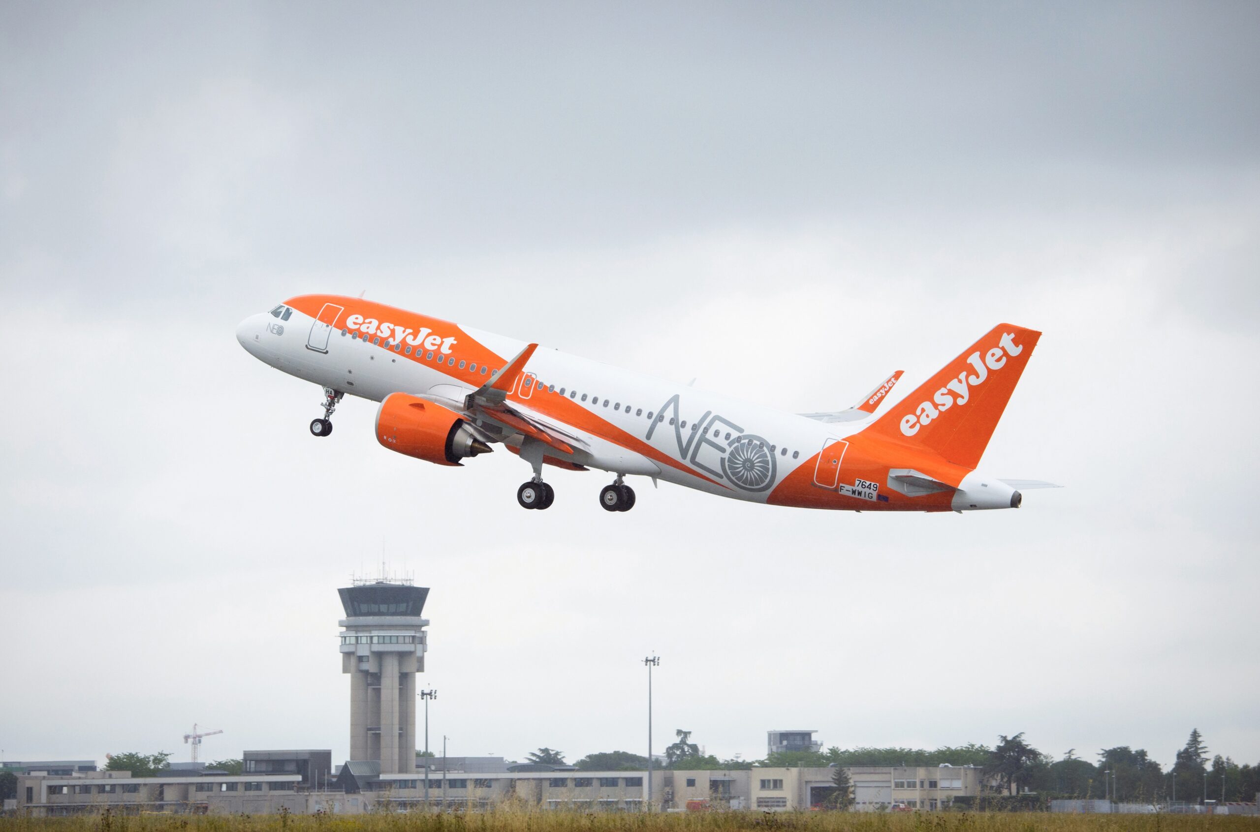 An orange and white EasyJet Airbus jet flying upwards over an airport, in front of a cloudy sky.