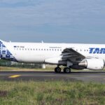 A blue and white Airbus jet in a TAROM livery. The jet is sat on a runway between two grassy fields.