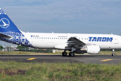 A blue and white Airbus jet in a TAROM livery. The jet is sat on a runway between two grassy fields.
