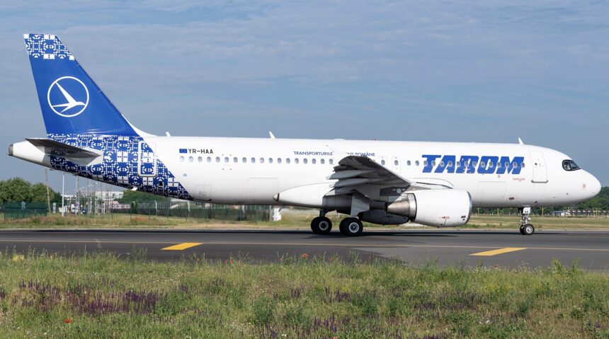A blue and white Airbus jet in a TAROM livery. The jet is sat on a runway between two grassy fields.