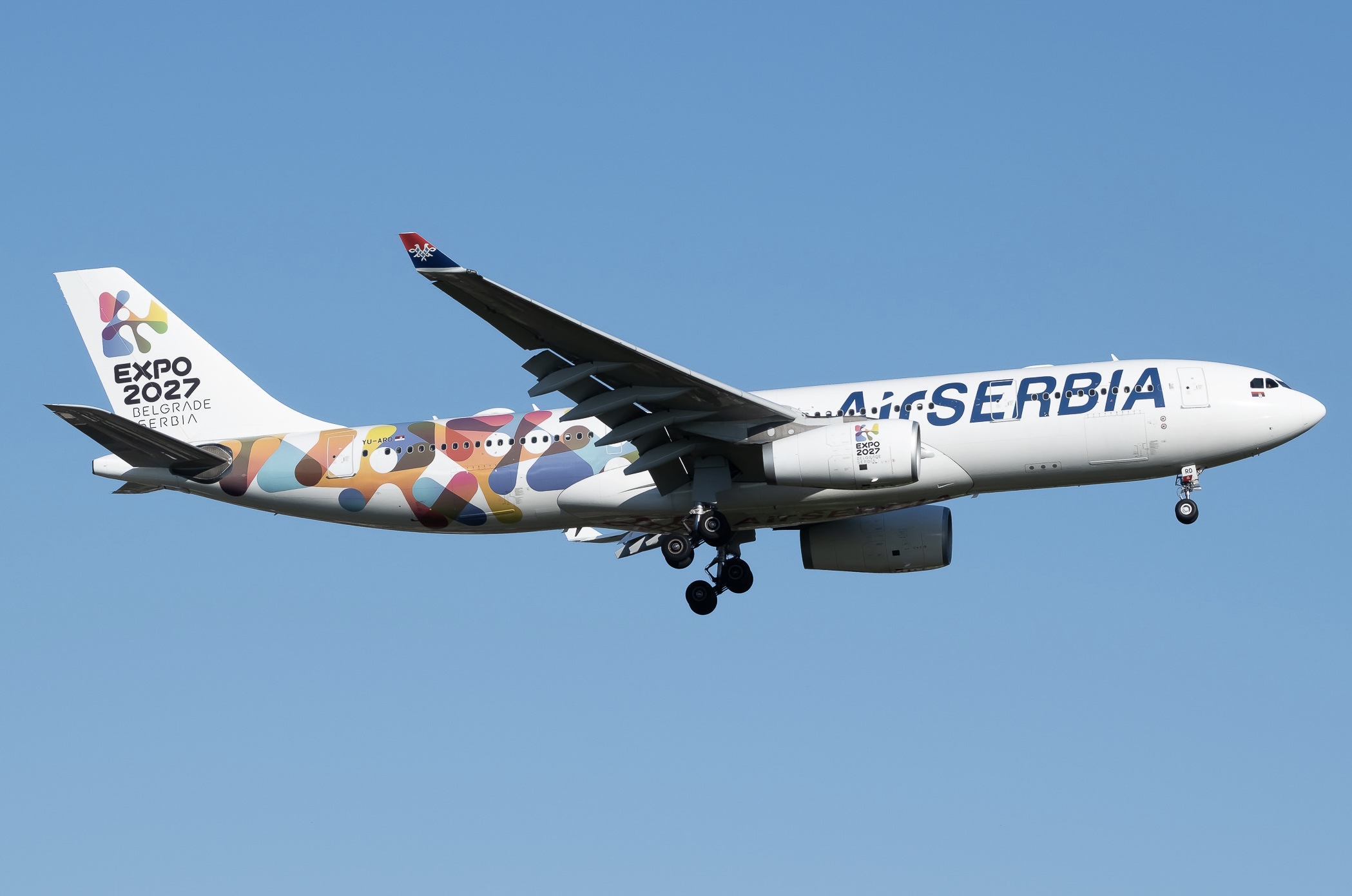 A white Air Serbia A330 jet with a multi-coloured tail flying in front of a clear blue sky.