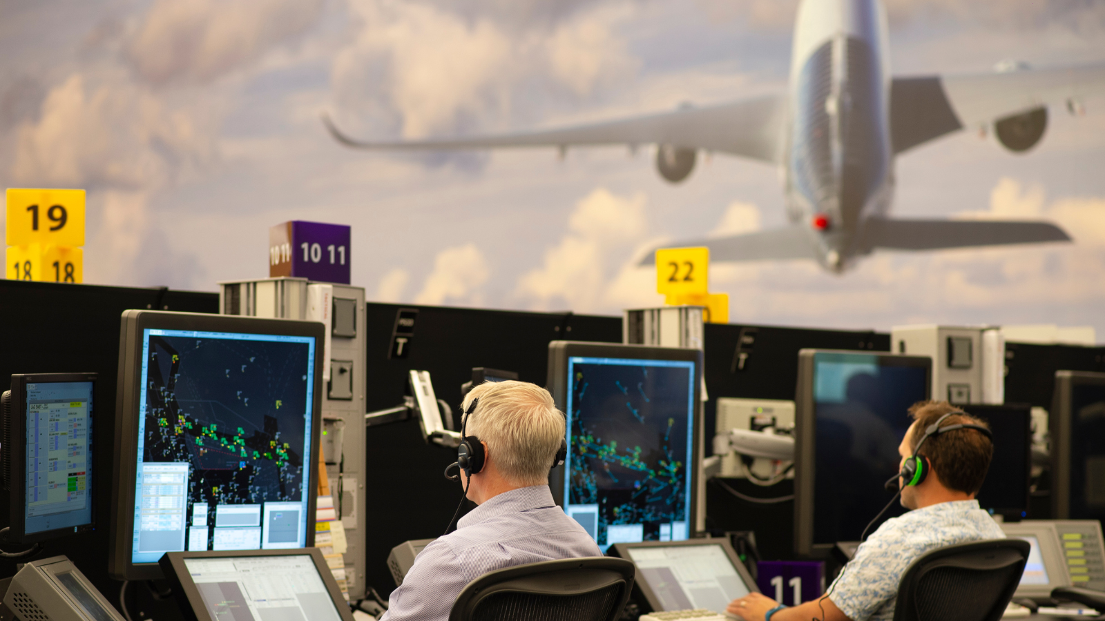 A team of air traffic controllers sat behind computer monitors. A picture of a jet flying over clouds is shown behind them.