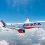 A red and white Air India jet flying over a layer of cloud in front of a blue sky.