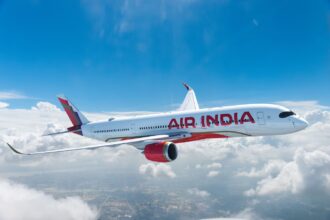 A red and white Air India jet flying over a layer of cloud in front of a blue sky.