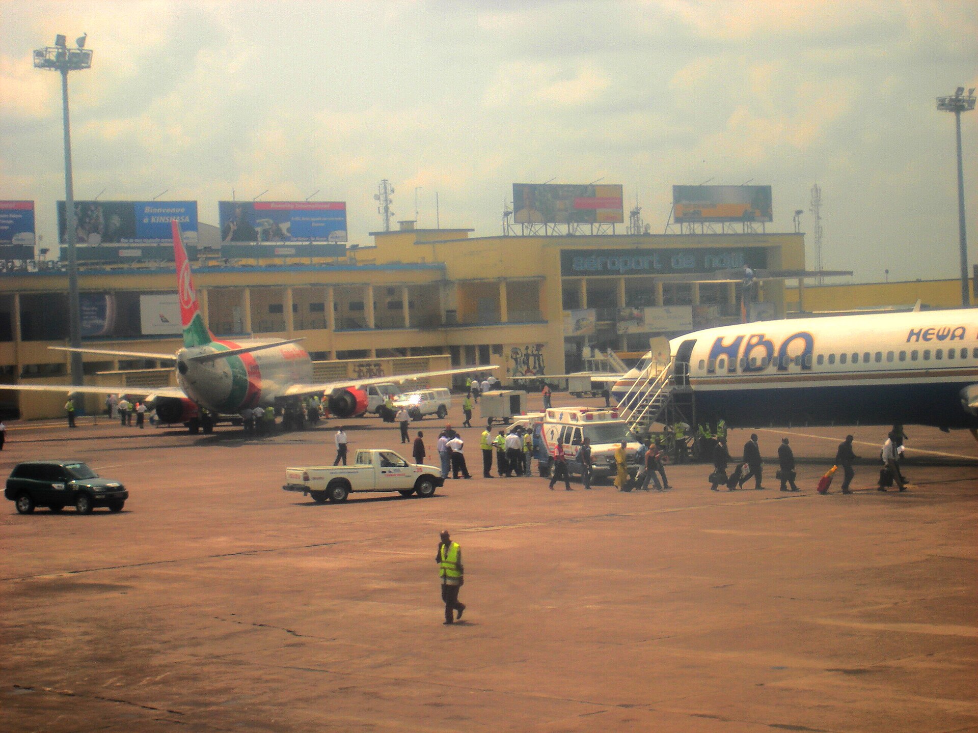 N’Djili International Airport (FIH). Two planes are sat in front of a terminal building which is shrouded by haze, beneath a cloudy sky.