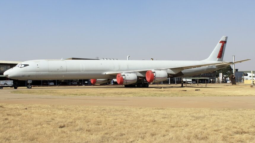 The red and white Douglas DC-8-73, in the livery of Trans Air Cargo Service. The plane is sat on a runway behind a field of yellow grass.