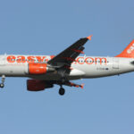 A white and orange easyJet Airbus A319-100 flying in front of a clear blue sky.