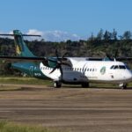 An ATR 72 jet in the green and white livery of Madagascar Airlines. The jet is sat on a runway in front of a small forest, on a clear day.