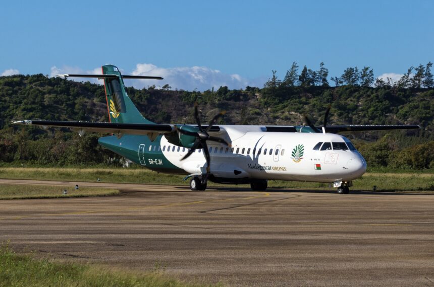 An ATR 72 jet in the green and white livery of Madagascar Airlines. The jet is sat on a runway in front of a small forest, on a clear day.