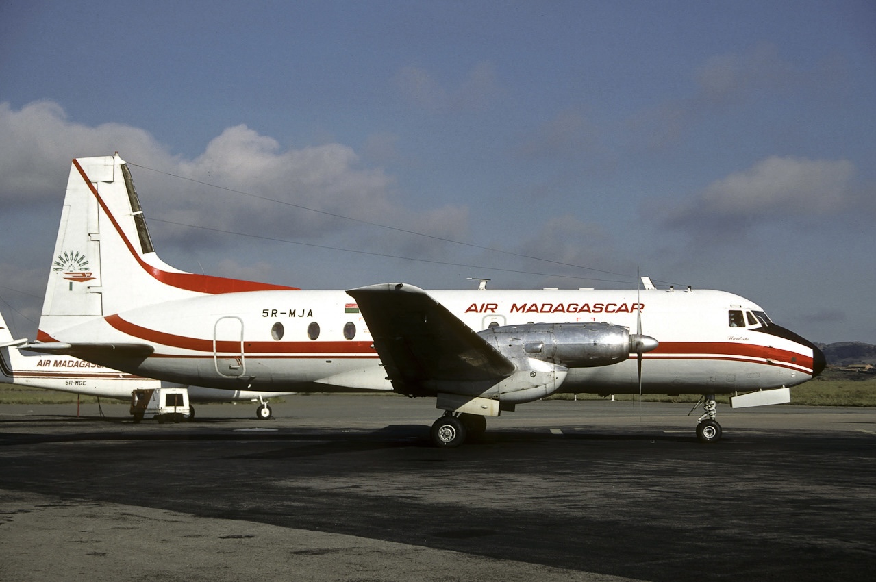 A white, red, and grey, Air Madagascar jet. The aircraft is sat on a runway in front of a cloudy sky.
