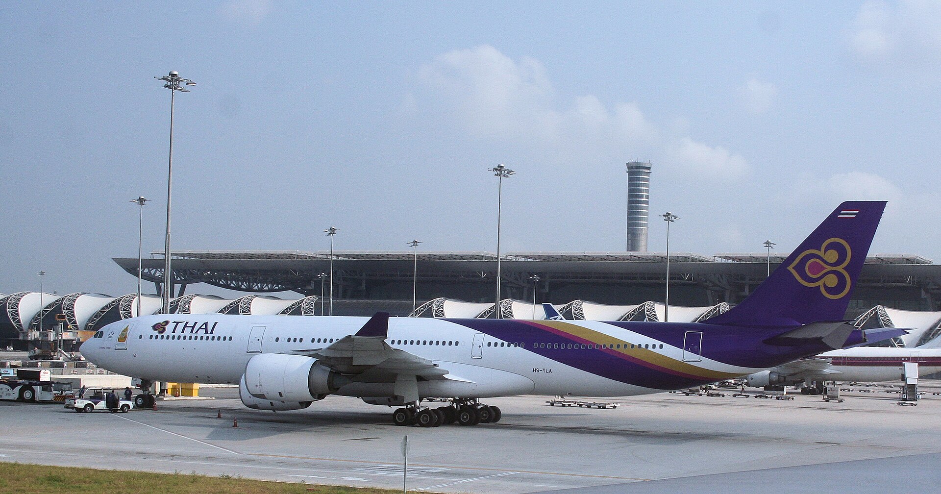 A white and purple Thai Airways International jet sat on a runway in front of a terminal building at Suvarnabhumi Airport (BKK) in Bangkok. The terminal is partially obscured by a heat haze.